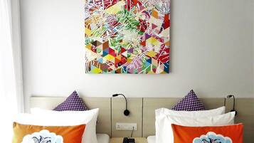 A hotel room with a bed, two pillows with floral designs, a bedside table with a phone, and a colorful abstract painting on the wall.