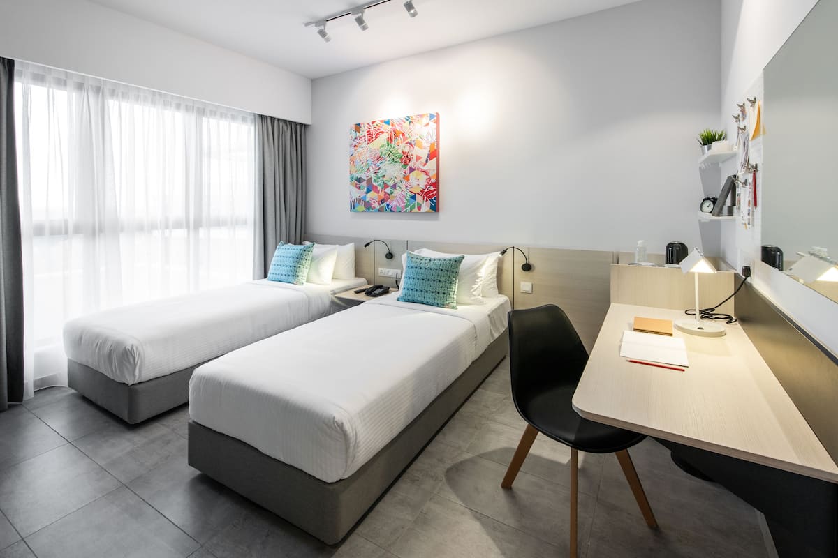 Quarto de hotel moderno com duas camas de solteiro, uma escrivaninha com cadeira, um espelho grande e uma pintura abstrata colorida na parede.
