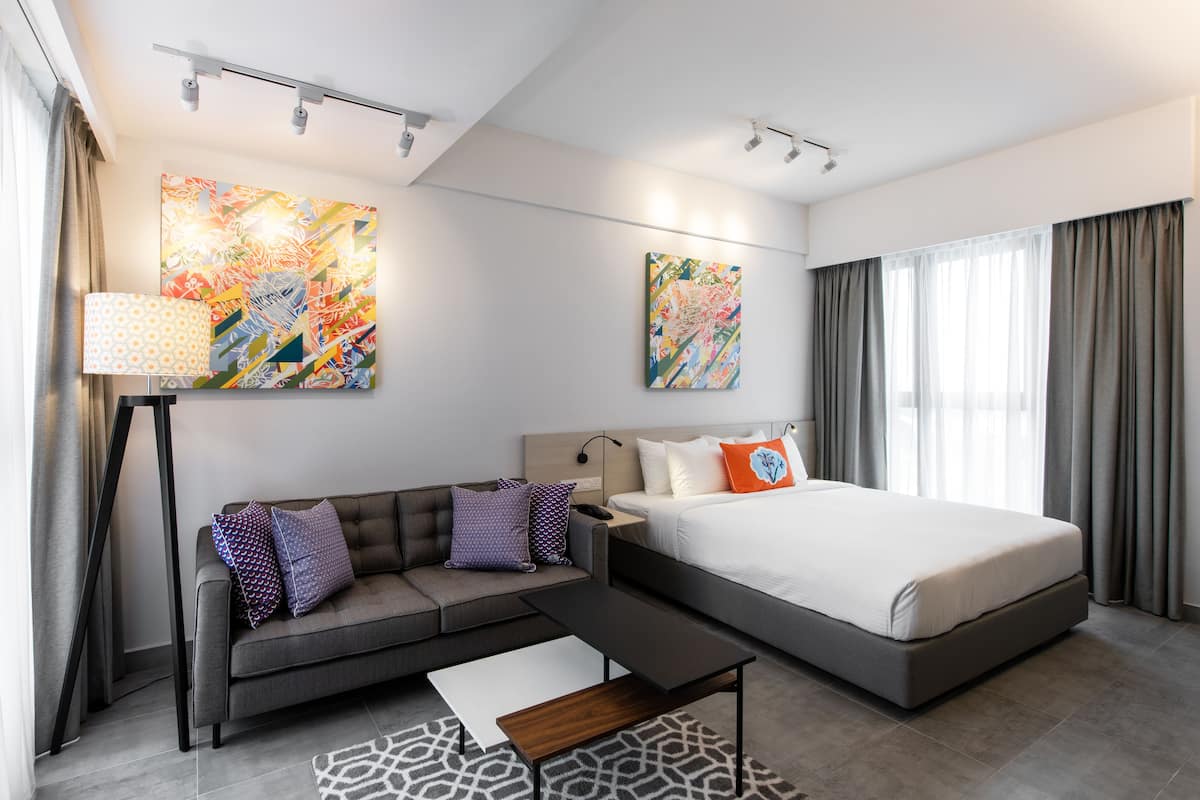 Quarto de hotel moderno com sofá, cama, escrivaninha e duas pinturas abstratas nas paredes.