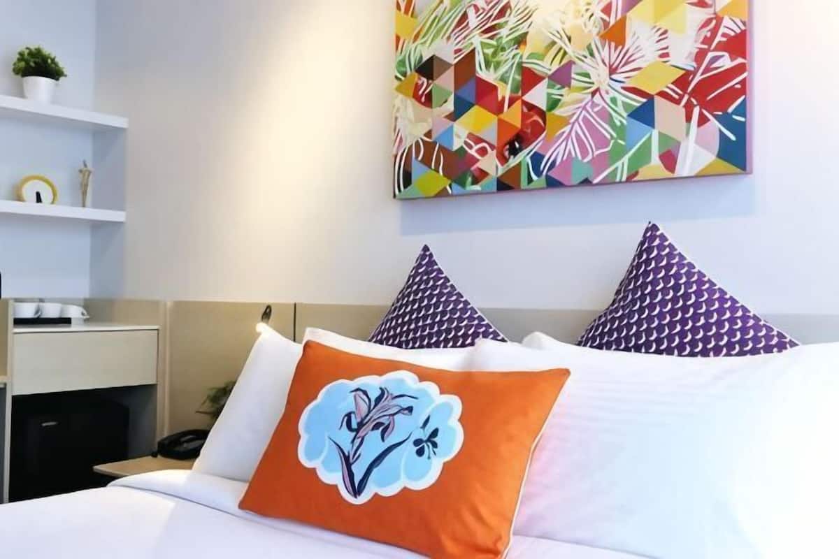 Quarto de hotel com cama, almofadas, uma obra de arte na parede e uma estante com objetos.
