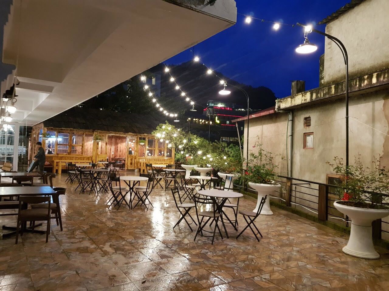 An outdoor restaurant area with tables and chairs, string lights, and a view of a building in the background.