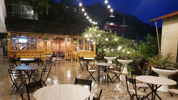 Un patio al aire libre con mesas y sillas redondas, una estructura de madera con ventanas y luces colgantes.