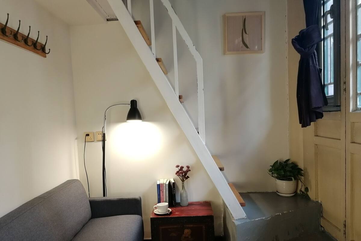 A living room with a sofa, a lamp, a small table with books and a vase, a staircase, and a potted plant.
