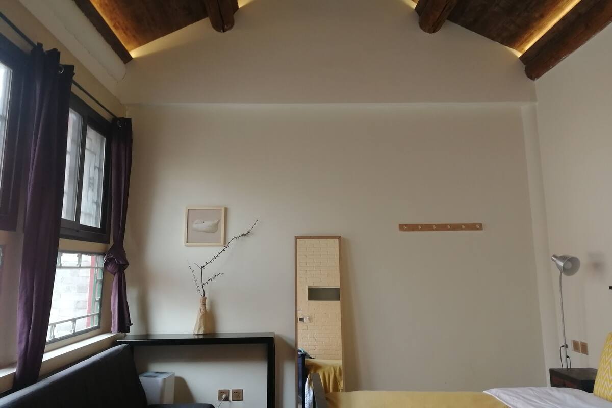 A room with a wooden ceiling, a bed, a desk with a chair, a window with curtains, a vase with branches, and a wall-mounted shelf.
