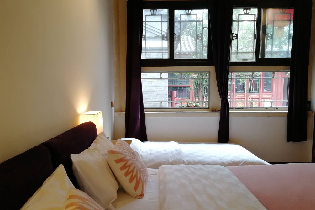 A hotel room with two beds, a window with curtains, and a view of buildings outside.