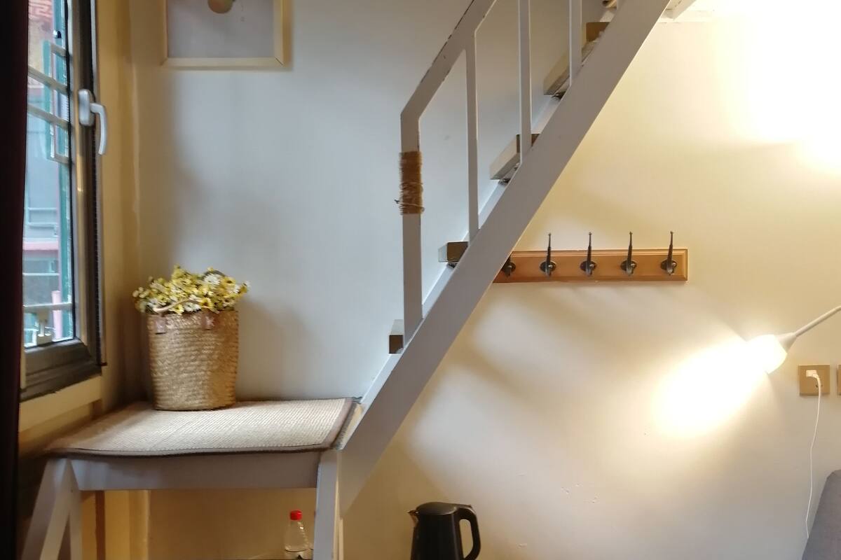 A staircase leading to a bedroom with a bed, a bedside table with a kettle, and a wall-mounted shelf with a basket of flowers.