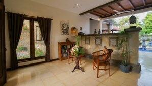A room with a tiled floor, a wooden chair, a table with a vase of flowers, a bookshelf, and a window with curtains. - Baliana Villa Batu Belig (Seminyak)