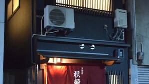 A building with a red awning displaying Japanese characters, an air conditioning unit on the roof, and a gas meter outside.
