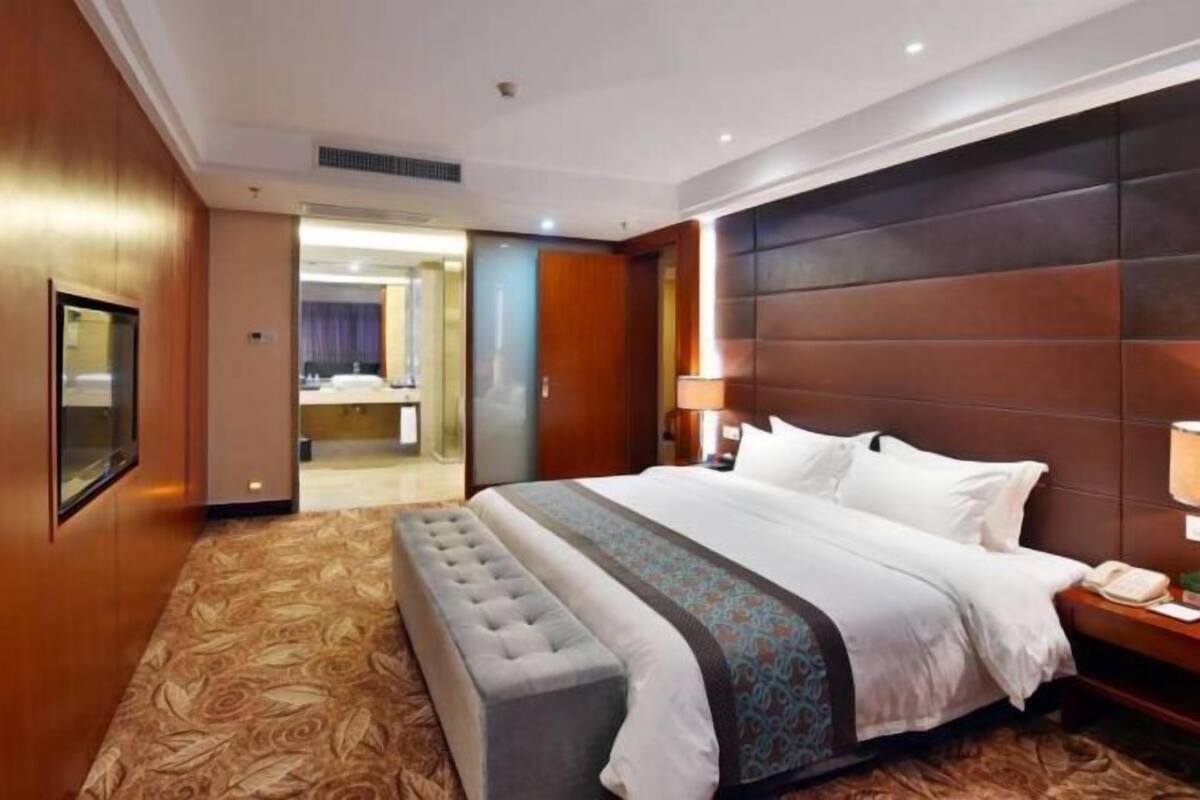 Quarto de hotel com uma cama grande, um banco, uma mesinha de cabeceira com abajur e um espelho.