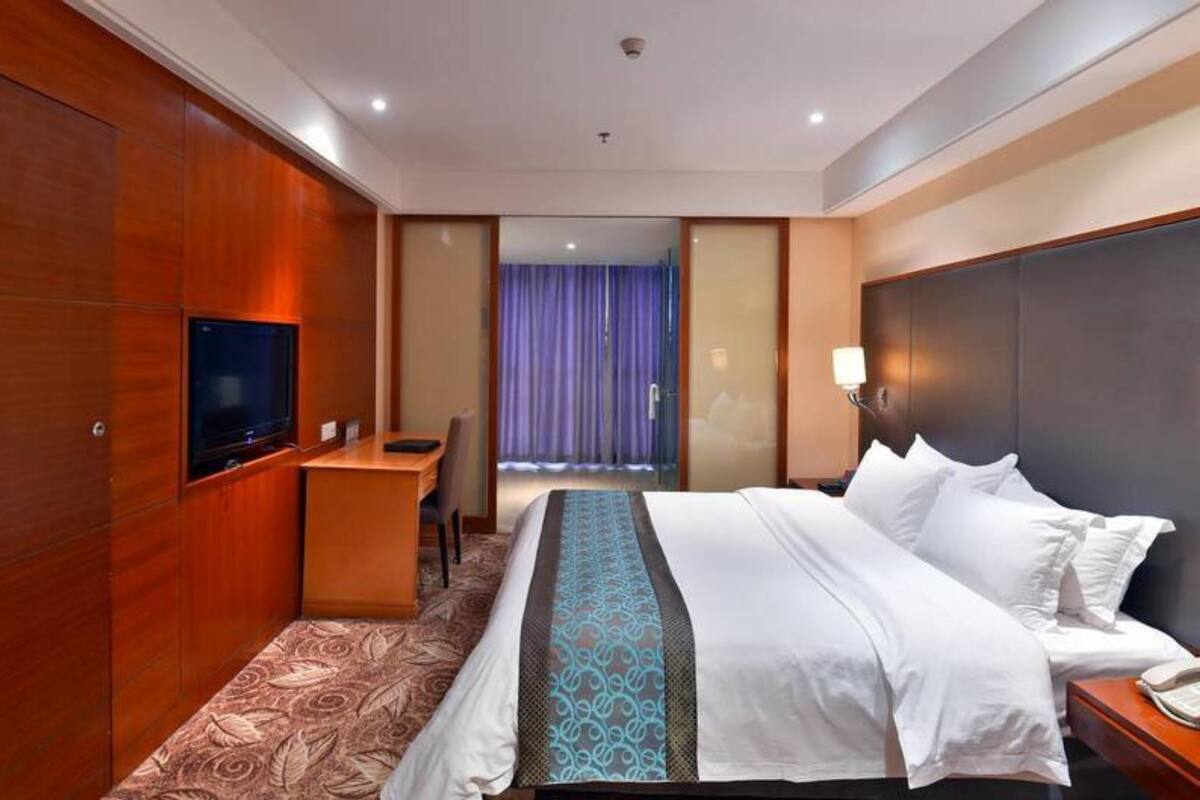 Quarto de hotel com uma cama grande, uma escrivaninha, uma televisão e vista para o exterior através de uma janela com cortinas.