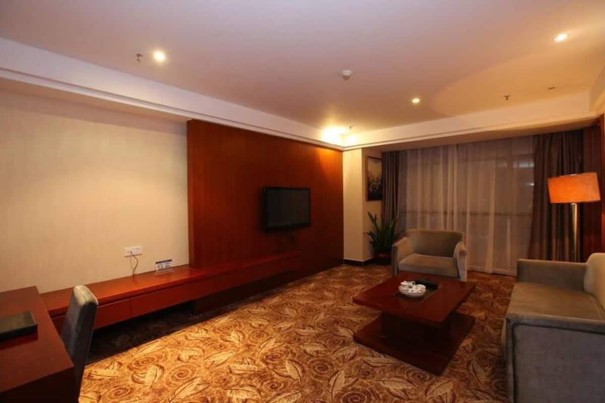 Quarto de hotel com uma cama grande, uma TV de tela plana na parede, uma área de estar com sofá e poltronas, e piso coberto por carpete.