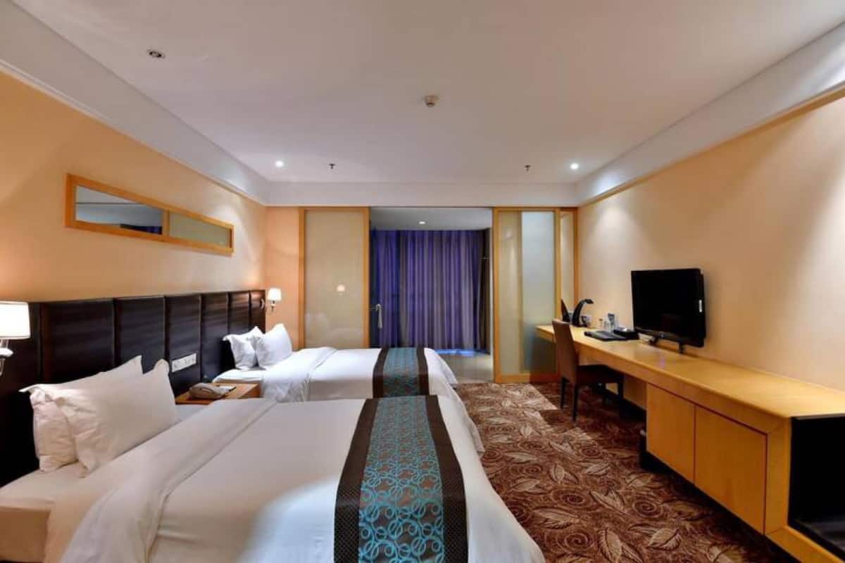 Quarto de hotel com duas camas, uma escrivaninha, televisão e banheiro visível por uma porta.