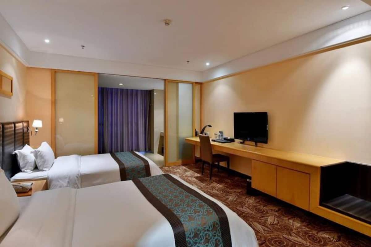 Quarto de hotel com duas camas, uma escrivaninha, televisão e banheiro com chuveiro.