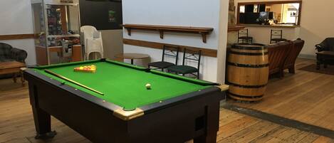 Una mesa de billar en una sala con piso de madera, una zona de bar con taburetes y un altavoz montado en la pared.