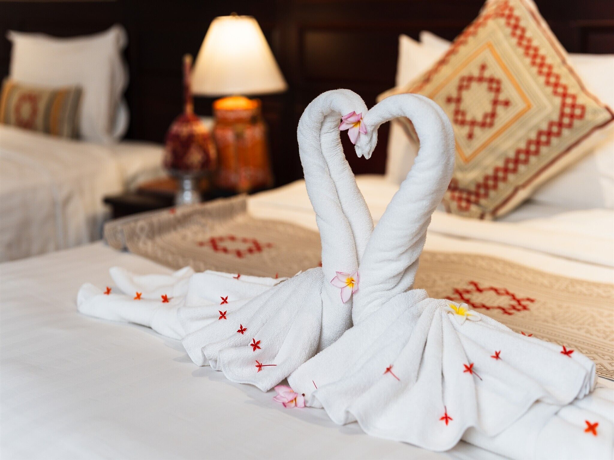 A bed with a towel arranged in the shape of two swans with hearts, a decorative pillow, and a lamp in the background.