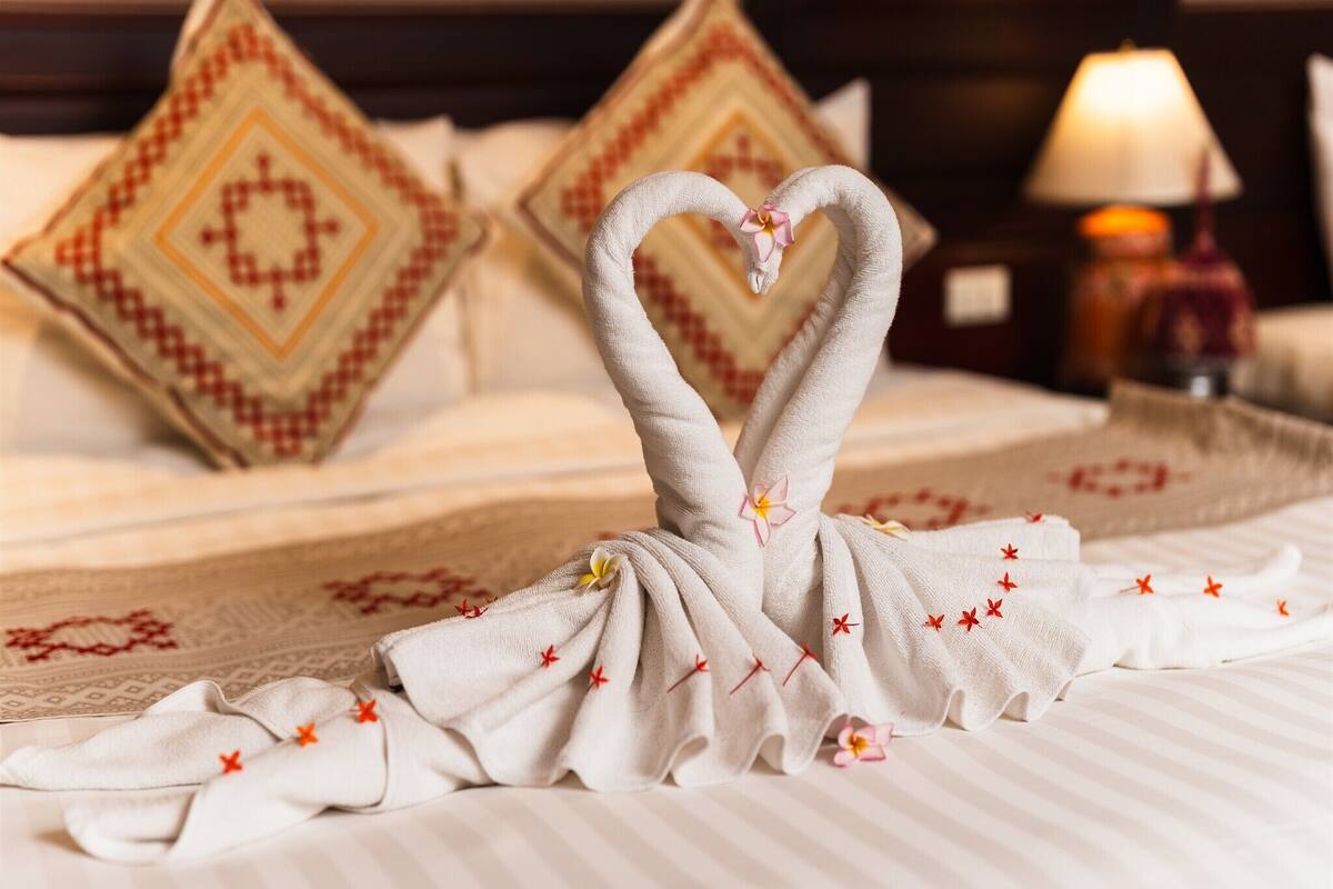 A neatly made bed with a towel arranged in a heart shape, pillows with a diamond pattern, and a lamp on the side.