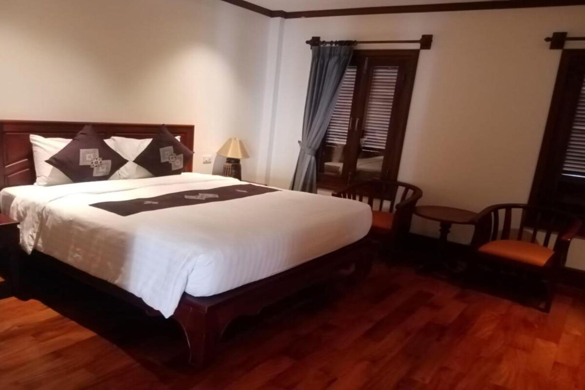 A hotel room with a wooden bed, a small table with a chair, and a window with blinds.