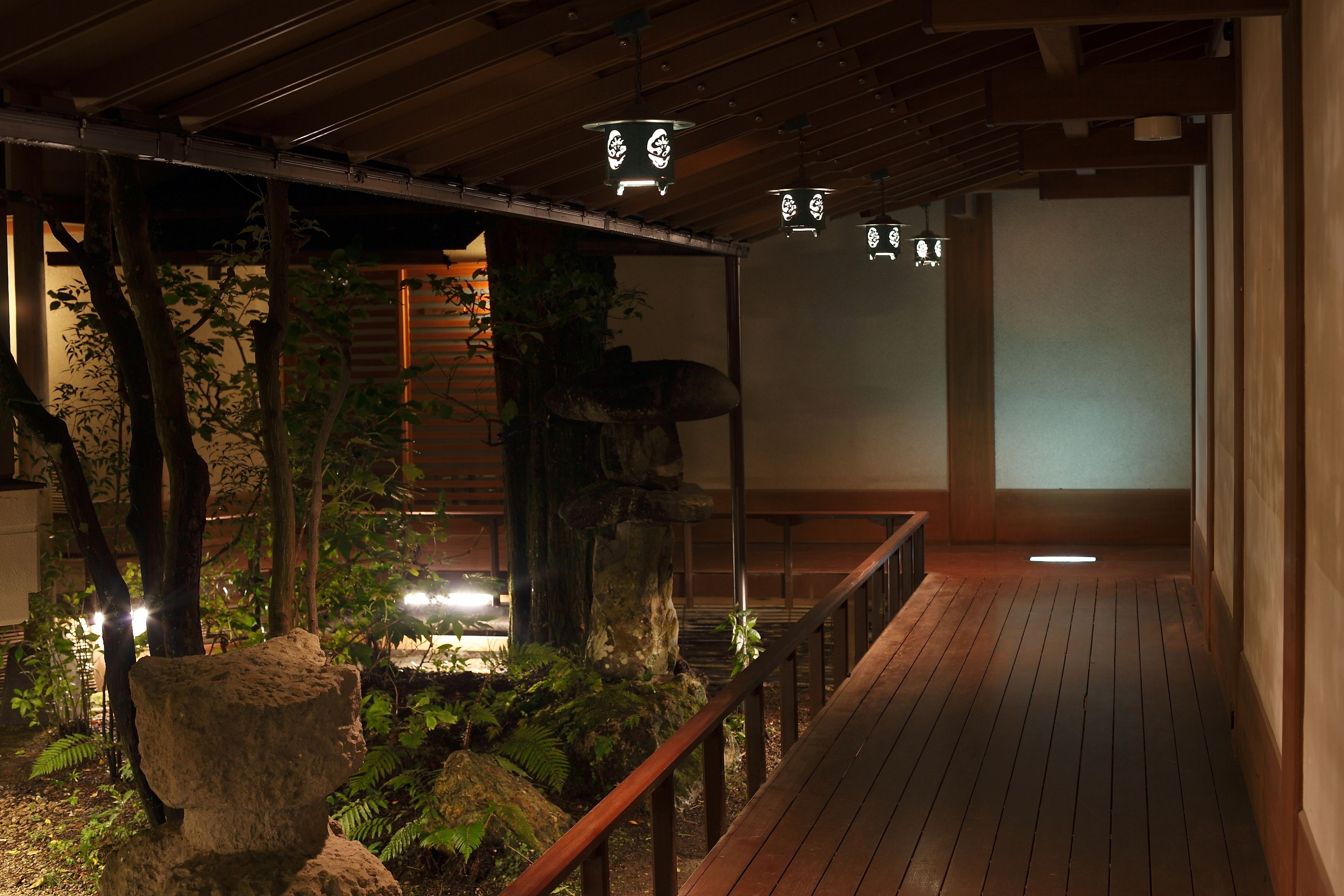 A wooden deck with a railing, surrounded by rocks and plants, leading to a traditional Japanese interior with tatami mats and shoji screens.