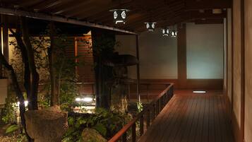 A wooden deck with a railing, surrounded by rocks and plants, leading to a traditional Japanese interior with tatami mats and shoji screens.