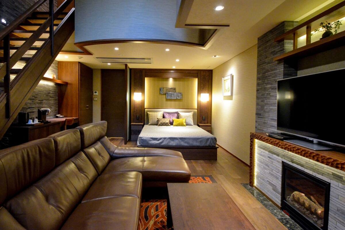 A modern hotel room with a curved staircase, a large bed with multiple pillows, a leather sofa, a flat-screen TV, and a fireplace.