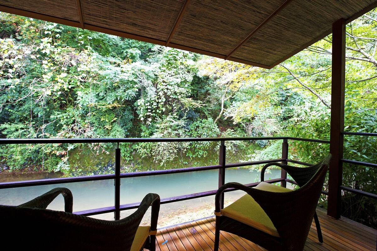 A wooden deck with two wicker chairs facing a river, surrounded by lush greenery.