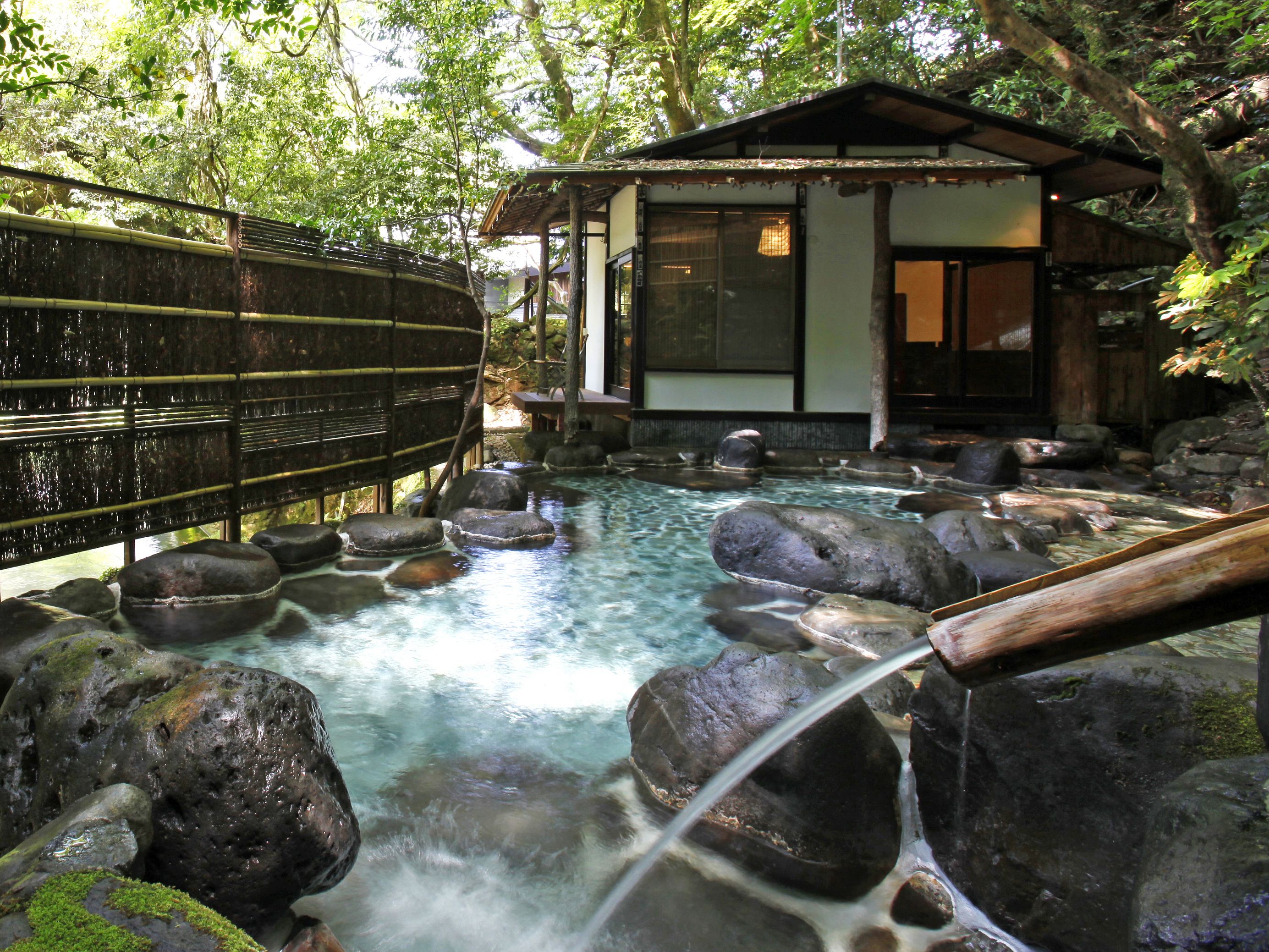 A traditional Japanese onsen with a wooden bathhouse, clear hot spring water, and a bamboo fence.