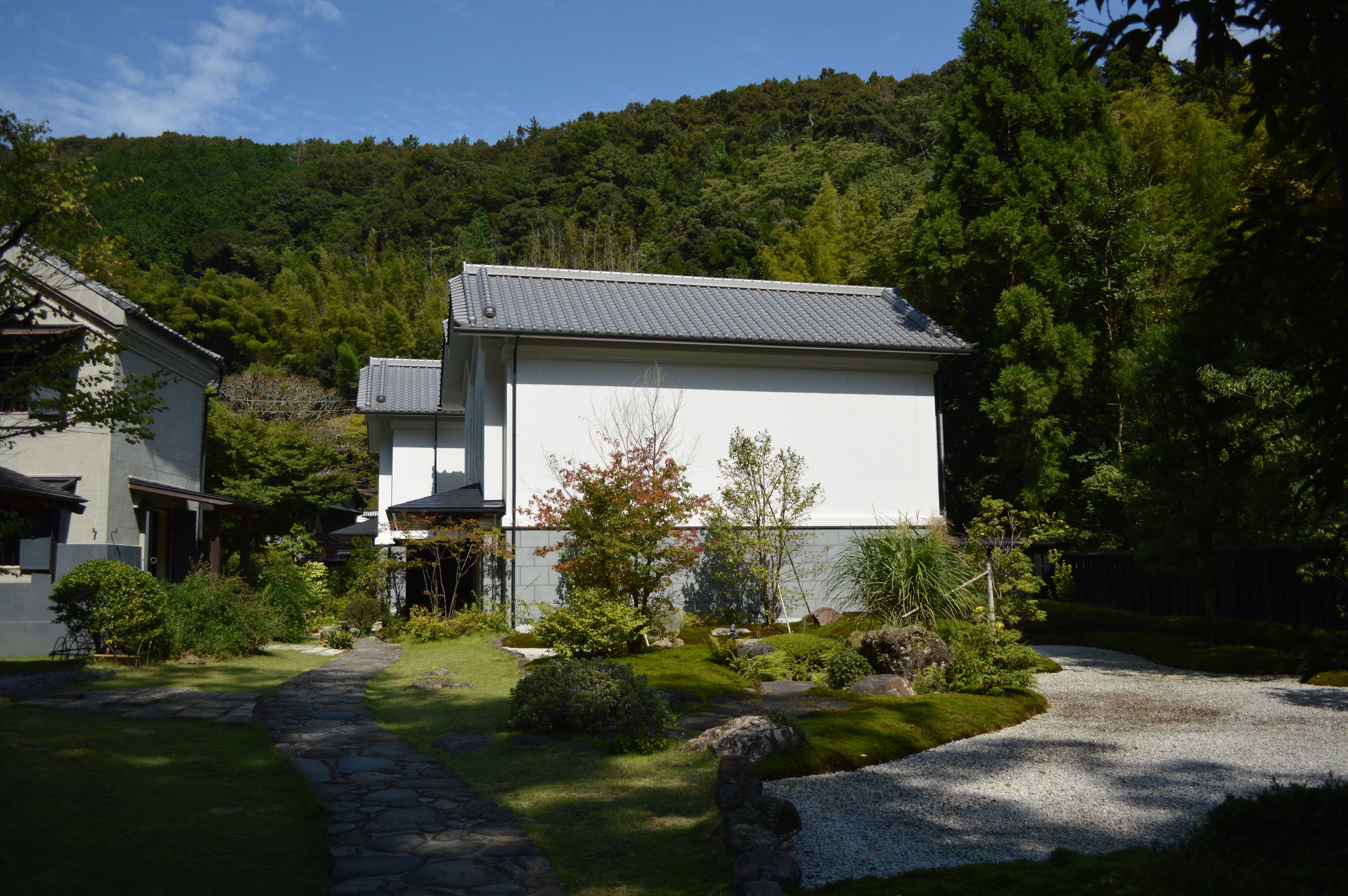 A modern house with a stone pathway leading to the entrance, surrounded by lush greenery and a forested hillside.