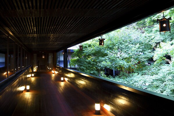 Un pasillo con suelo de madera y faroles colgantes, que conduce a un muro de vidrio con vistas a un jardín exuberante.