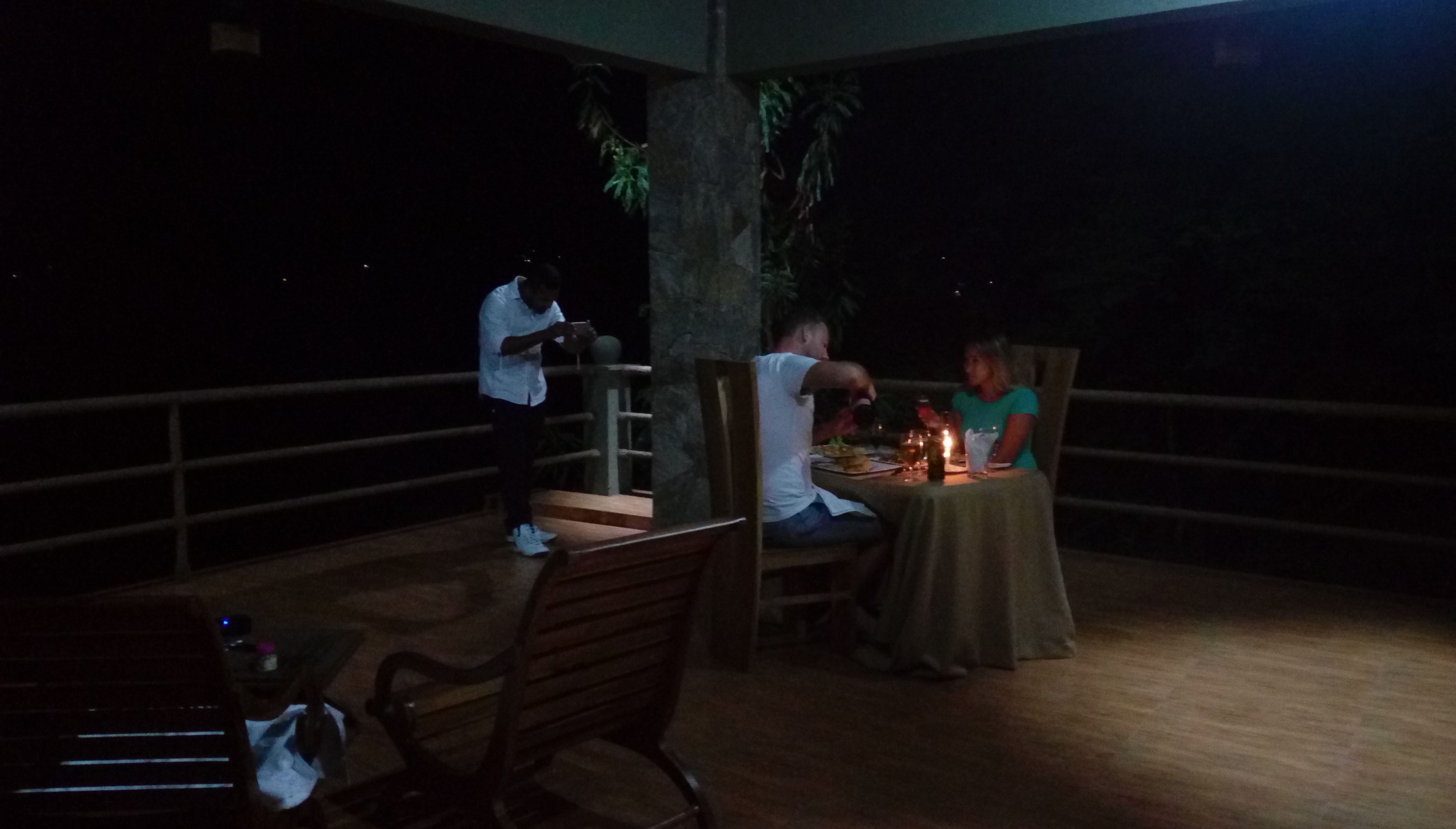 An outdoor dining area with a table and chairs, a person standing, and another person seated at the table.