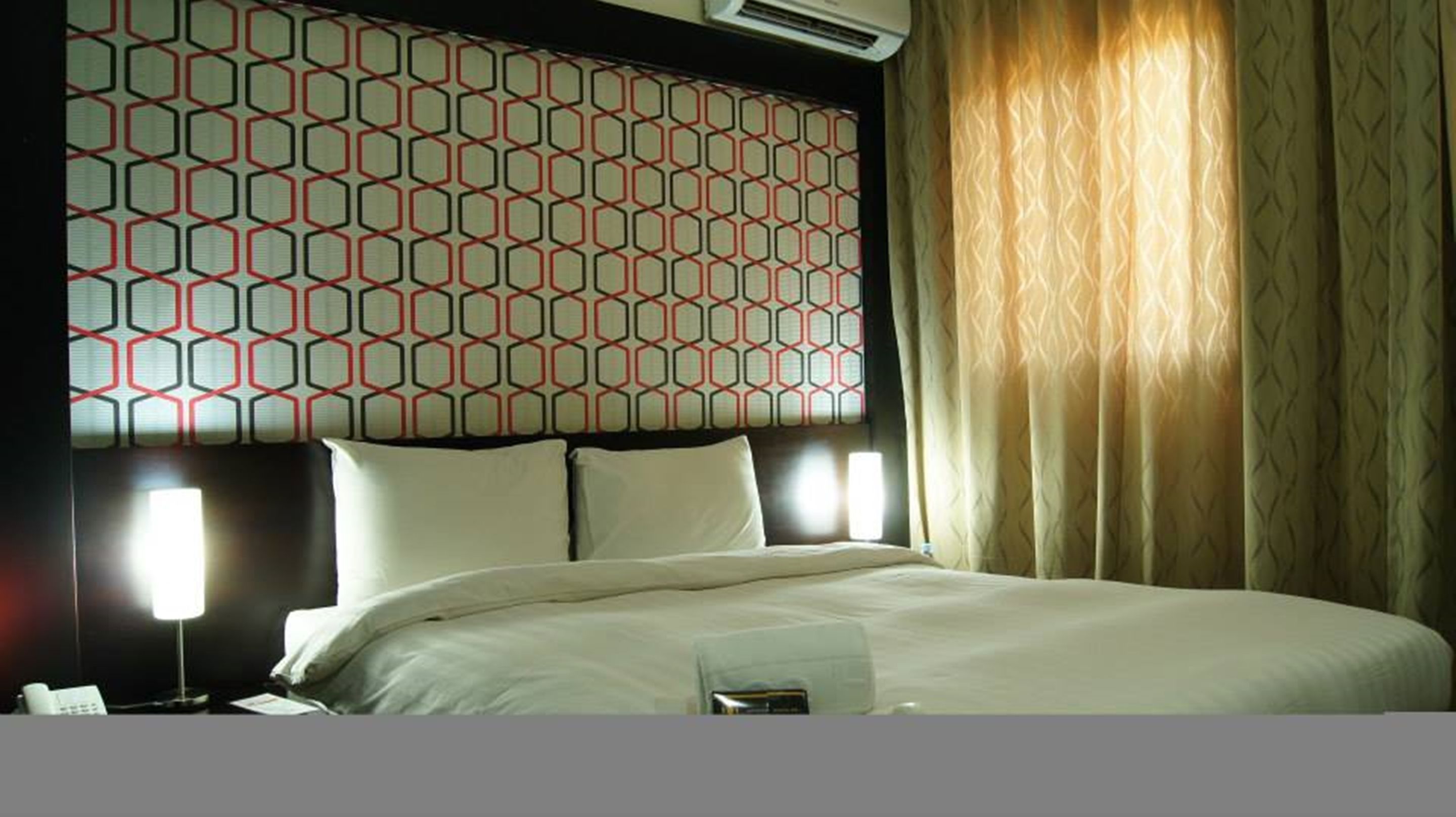 A hotel room with a patterned headboard, a bed with white linens, two bedside lamps, and a window with curtains.