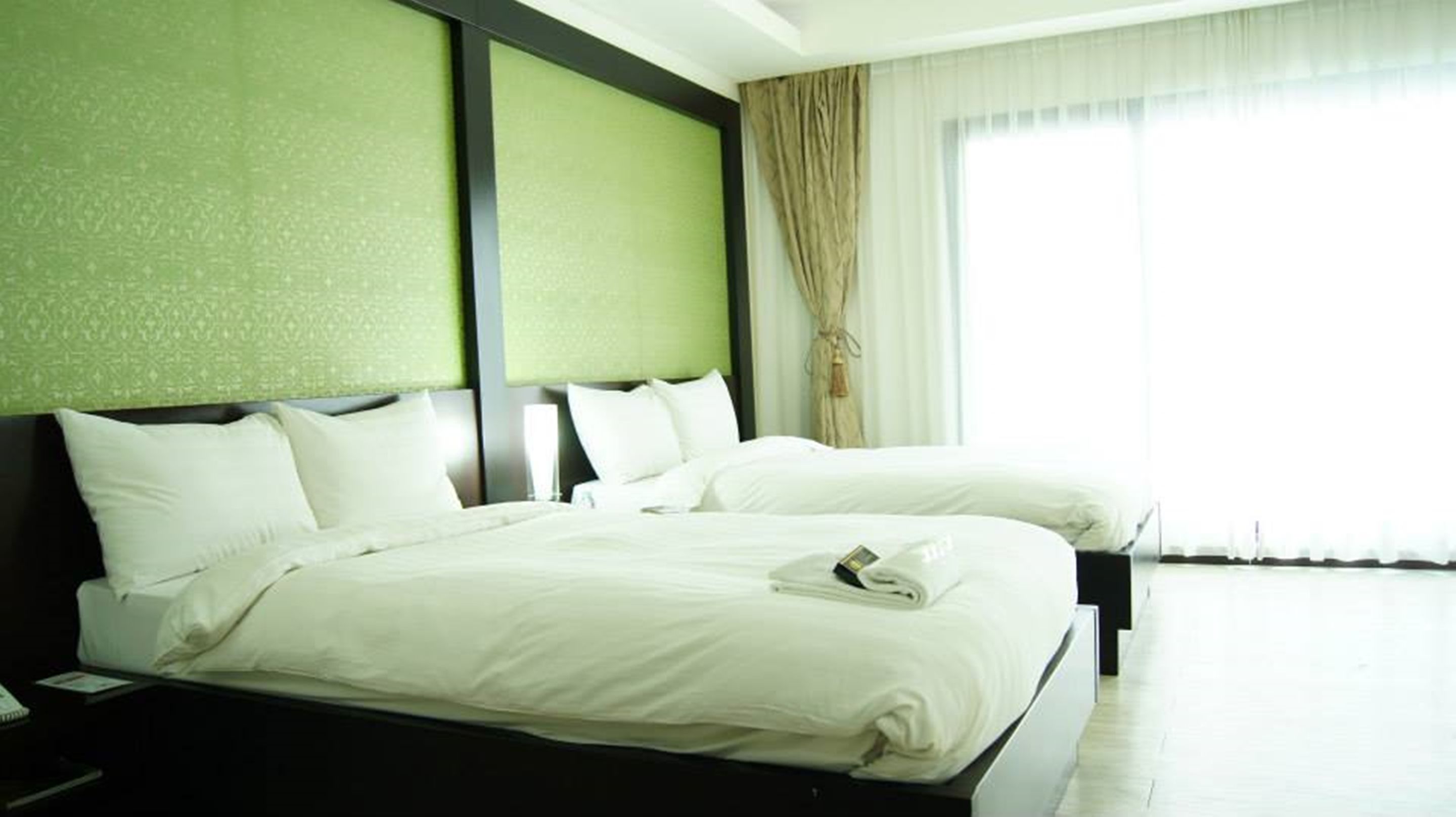 A hotel room with two beds, a green headboard, white bedding, and a large window with curtains.