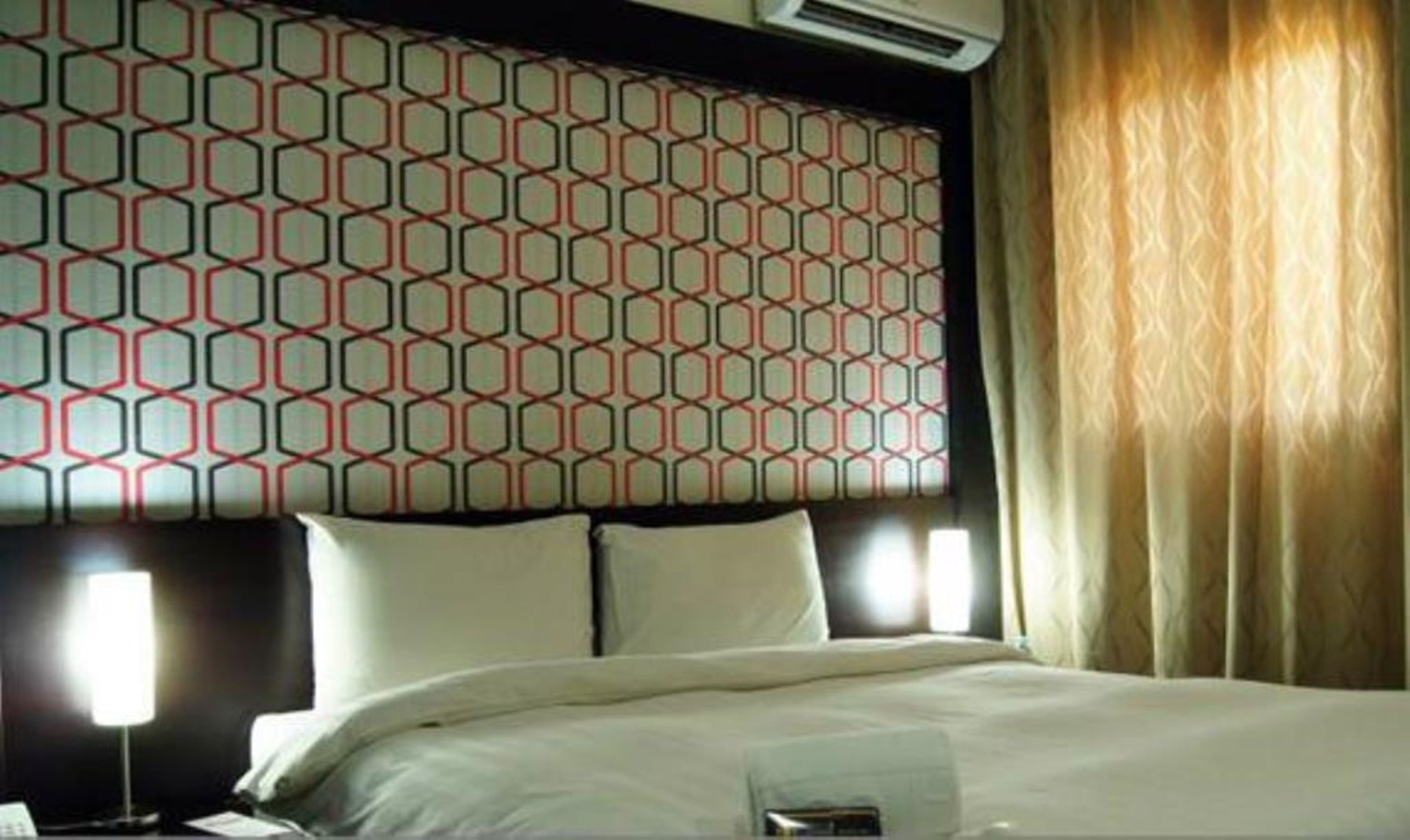 Une chambre d’hôtel avec une tête de lit à motifs, un lit avec du linge de maison blanc, une lampe de chevet et une fenêtre avec des rideaux.