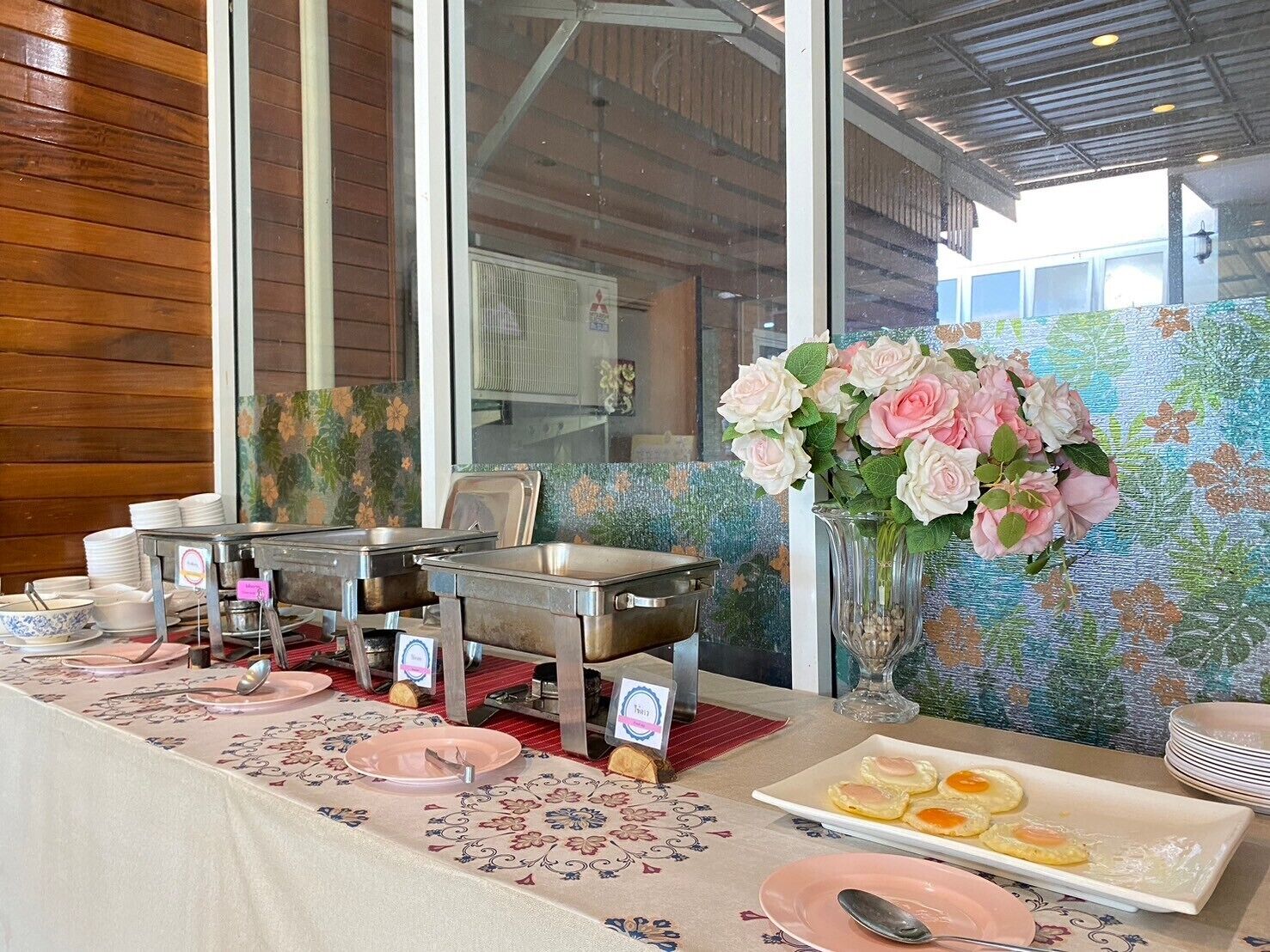 A breakfast buffet setup with stainless steel chafing dishes, a floral centerpiece, and a table with pink plates and a white plate with eggs.
