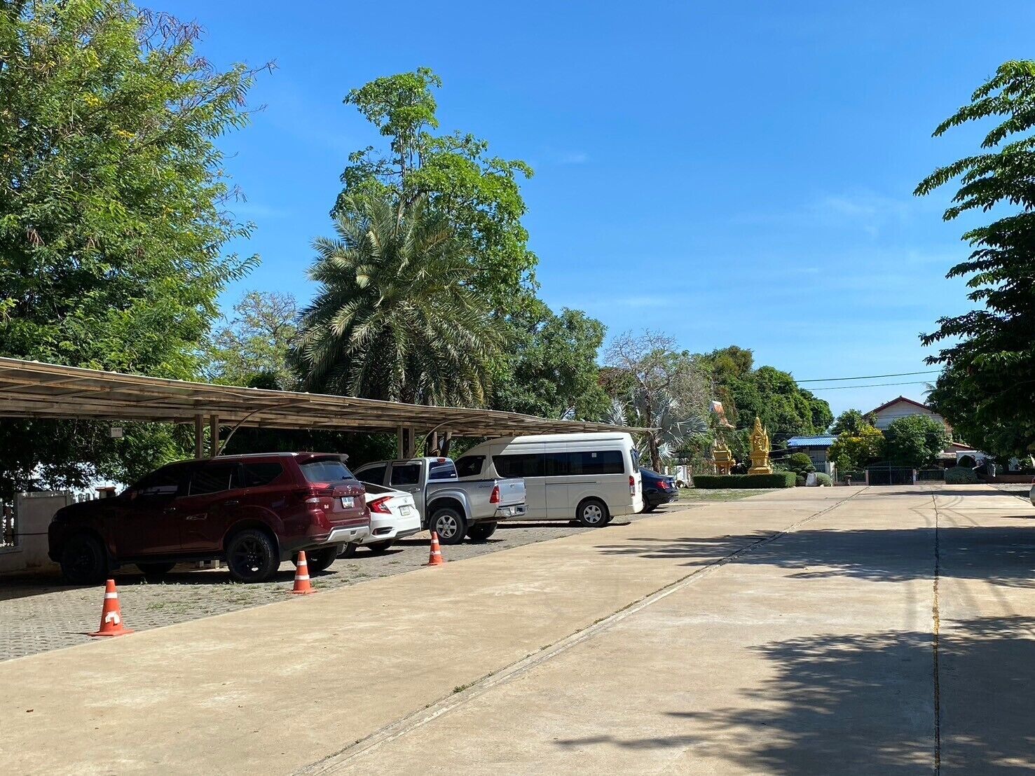 A parking area with several vehicles, including SUVs and a van, under a covered structure with trees and a building in the background.