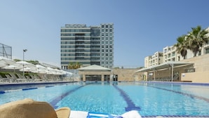 A swimming pool with lounge chairs and umbrellas, a tall building in the background, and a hat and goggles on the poolside. - Okeanos ba Marina by Sophie (Herzliya)
