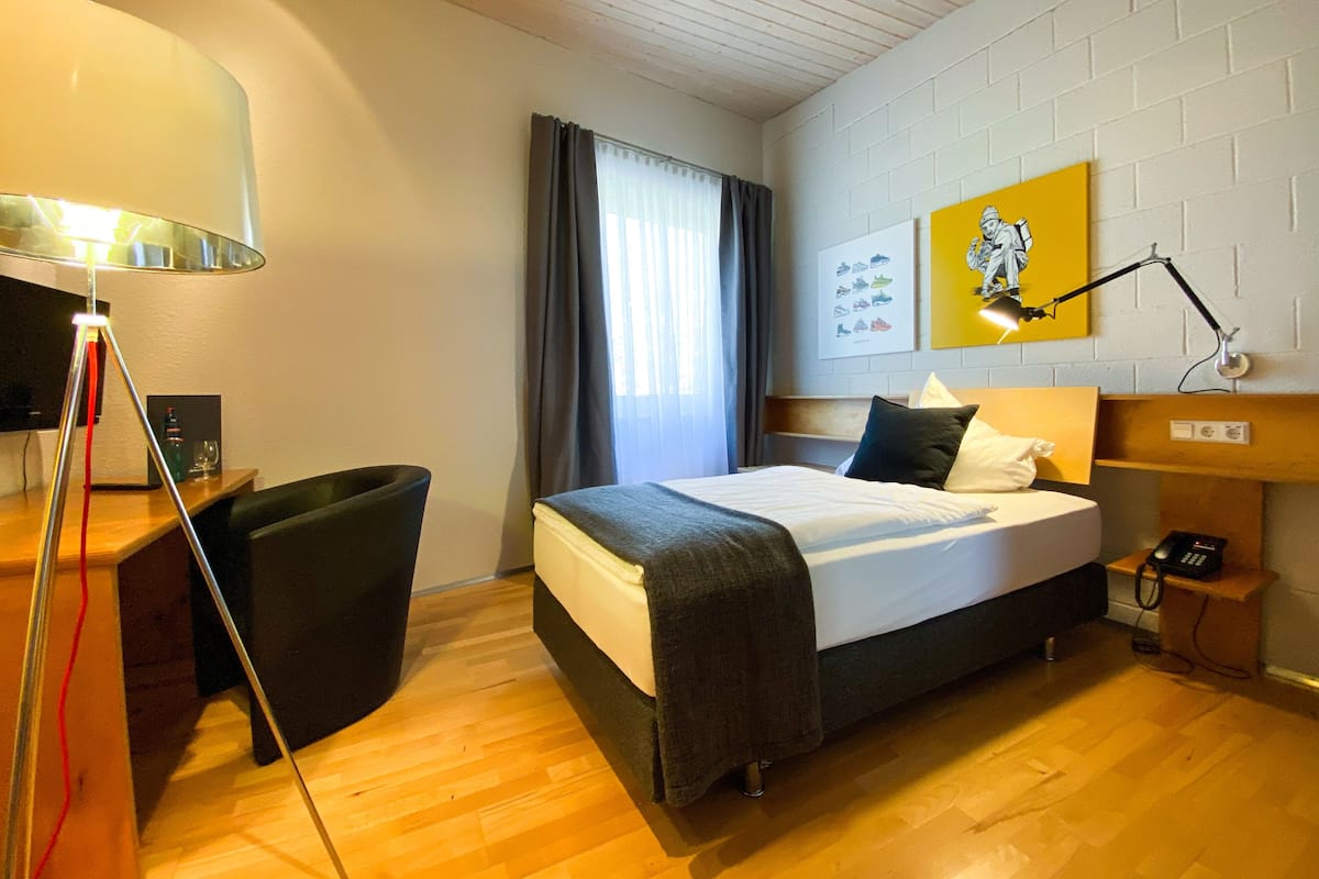 A hotel room with a bed, a desk, a chair, a lamp, a painting on the wall, and a window with curtains.