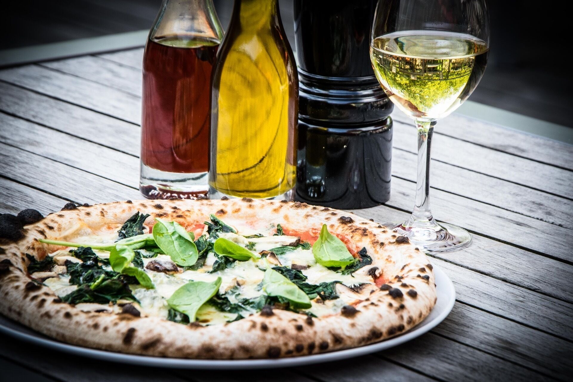 A pizza with spinach and tomato on a wooden table, accompanied by a wine glass and two bottles of olive oil.