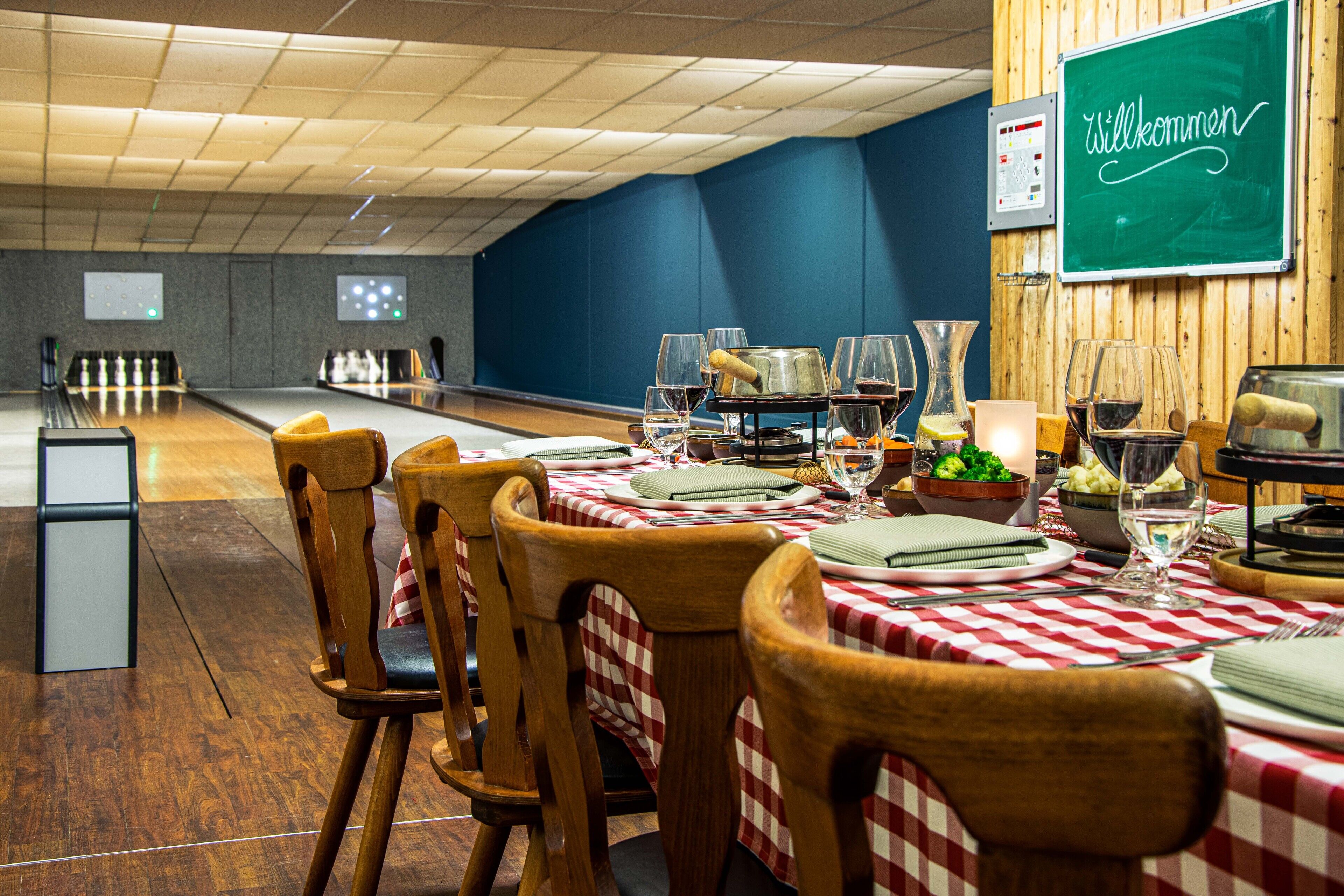 A restaurant with wooden chairs and tables, a chalkboard with "Willkommen" written on it, and bowling lanes in the background.