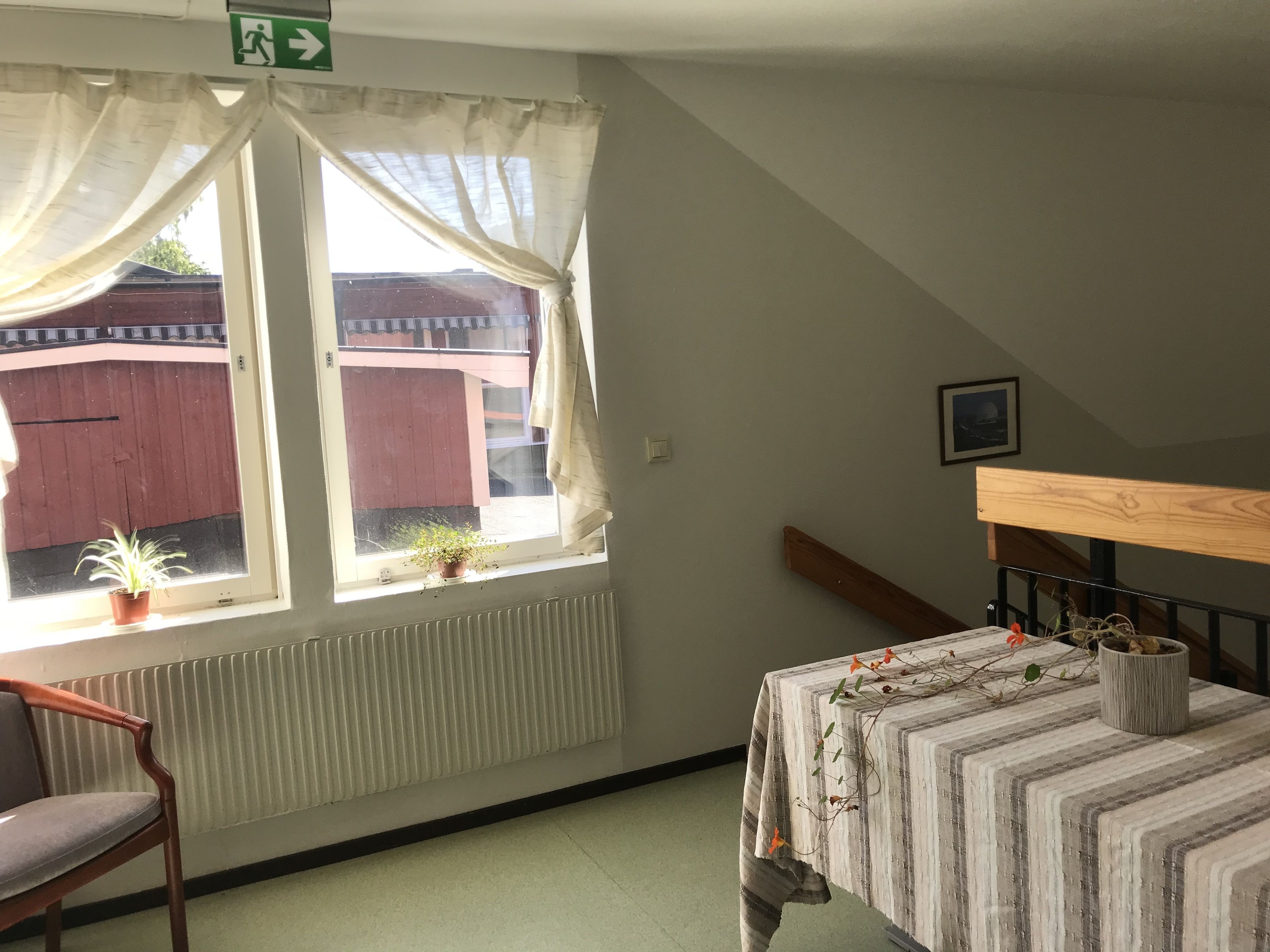 A room with a window, a radiator, a chair, a table with a striped tablecloth, and a wooden headboard.