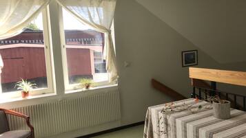 A room with a window, a radiator, a chair, a table with a striped tablecloth, and a wooden headboard.