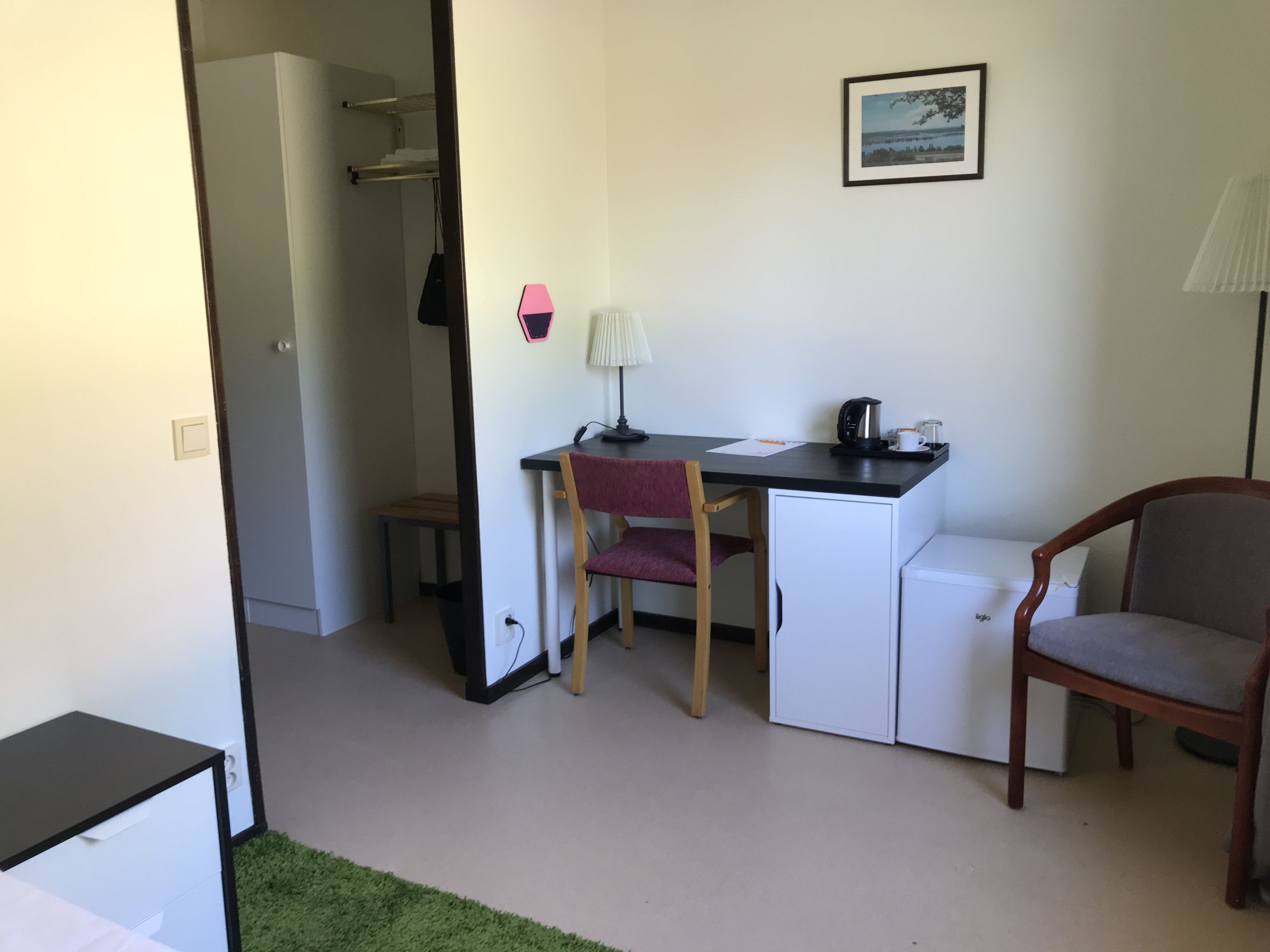 Habitació compacta amb escriptori, cadira i un petit frigorífic. Hi ha una perxa i una làmpada a la paret.