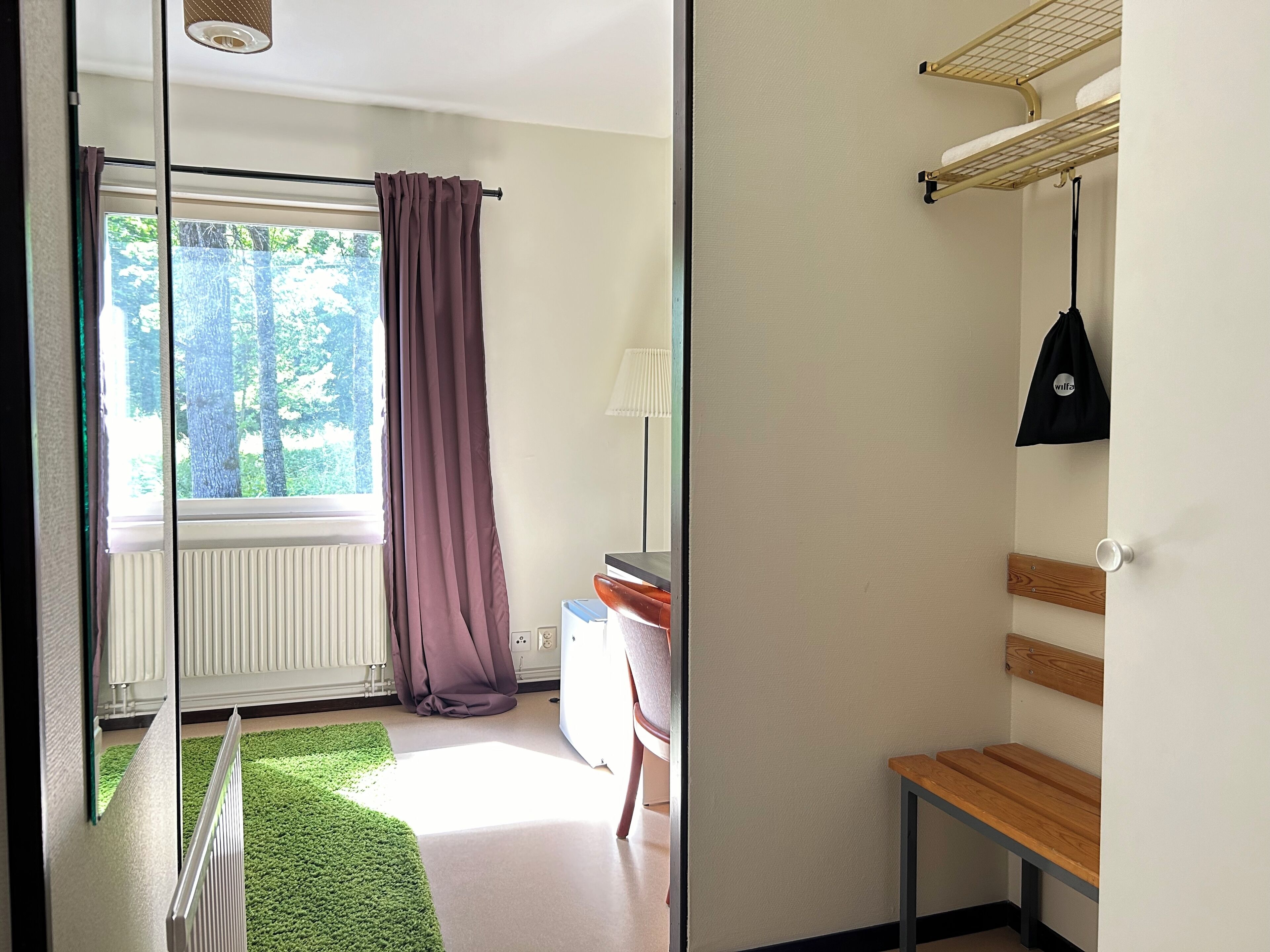 Quarto com janela, radiador, tapete verde, banco de madeira e prateleira fixada na parede com uma bolsa.
