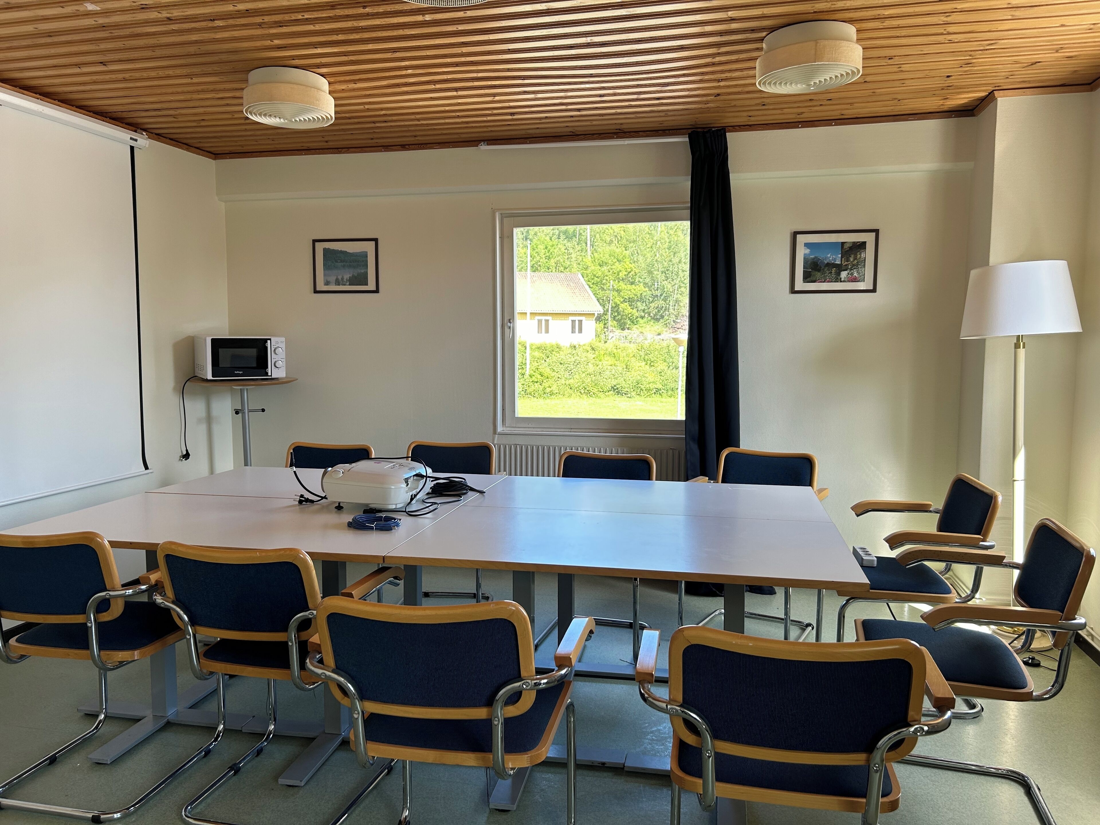 A conference room with a long table, six chairs, a projector, and a window with a view of greenery.