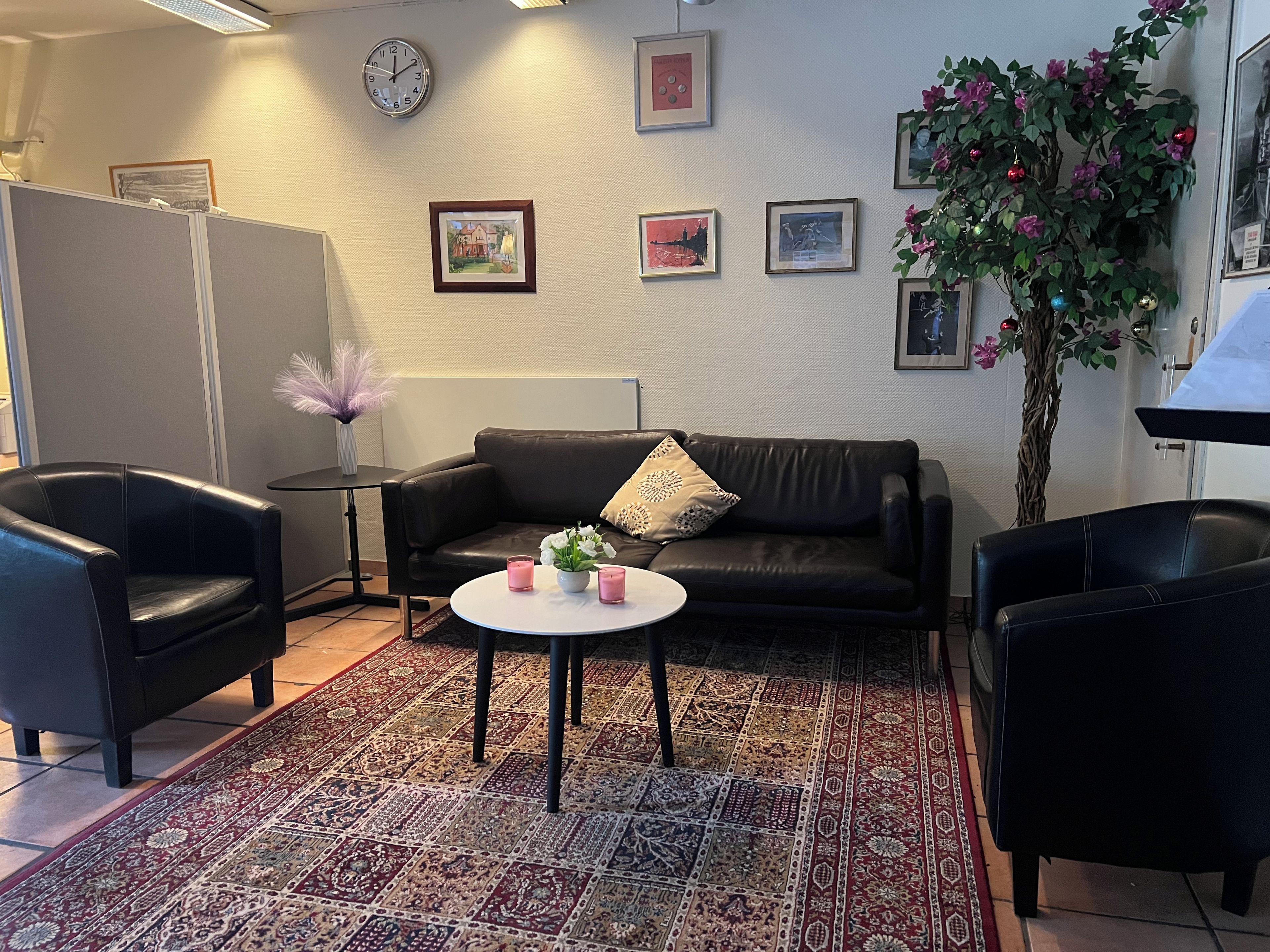 Un salon avec un canapé en cuir noir, deux fauteuils noirs, une petite table blanche ornée de bougies roses, un tapis à motifs et un mur décoré de tableaux encadrés.