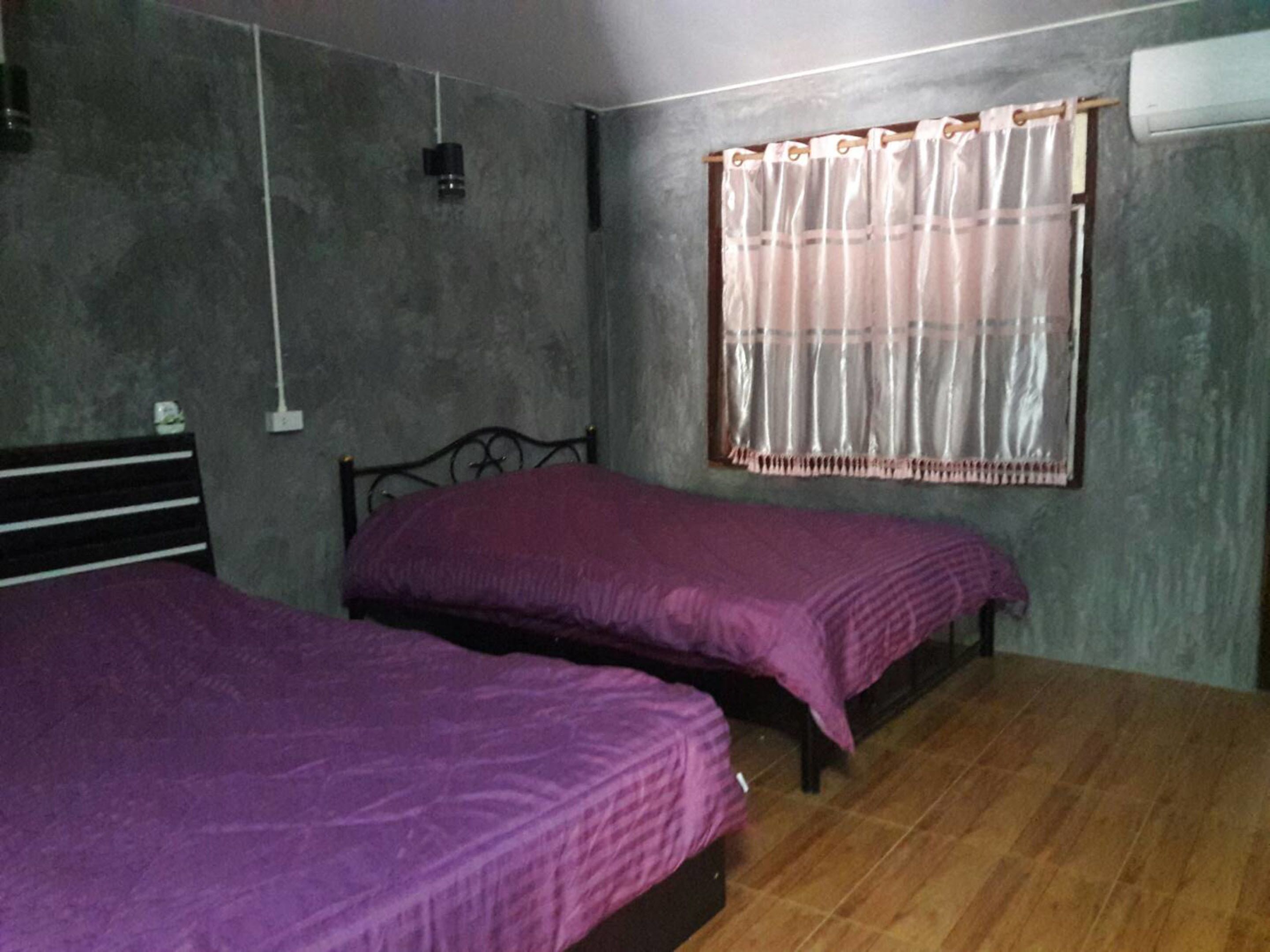 A room with two beds, a window with curtains, a wall-mounted air conditioner, and a wooden floor.