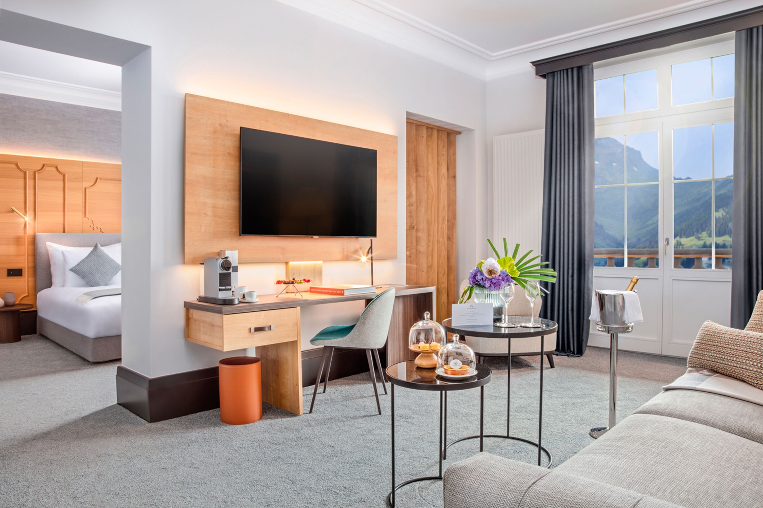 Una habitación de hotel moderna con una cama grande, un escritorio con televisión de pantalla plana, una mesita con un jarrón y vistas a las montañas a través de la ventana.
