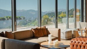 A modern lounge with a sofa, ottoman, and round table, featuring a mountain view through large windows. - Villars Palace (Ollon)