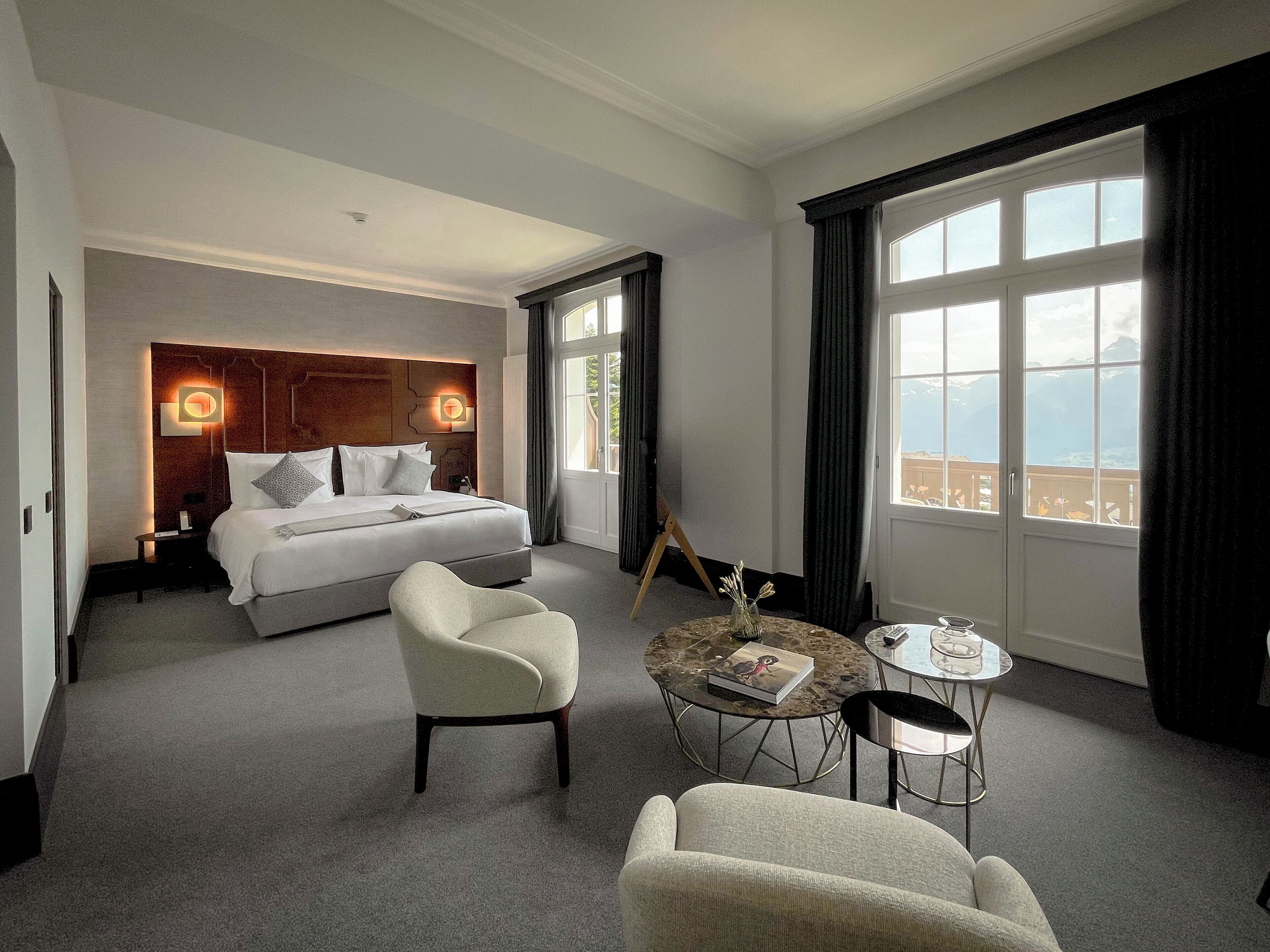 Habitación de hotel con una cama grande, dos sillones, una mesita redonda y vistas a las montañas a través de las ventanas.