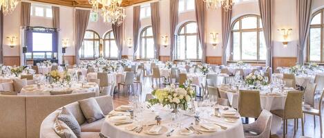 Un amplio salón de banquetes con mesas redondas, elegantes candelabros y grandes ventanales.