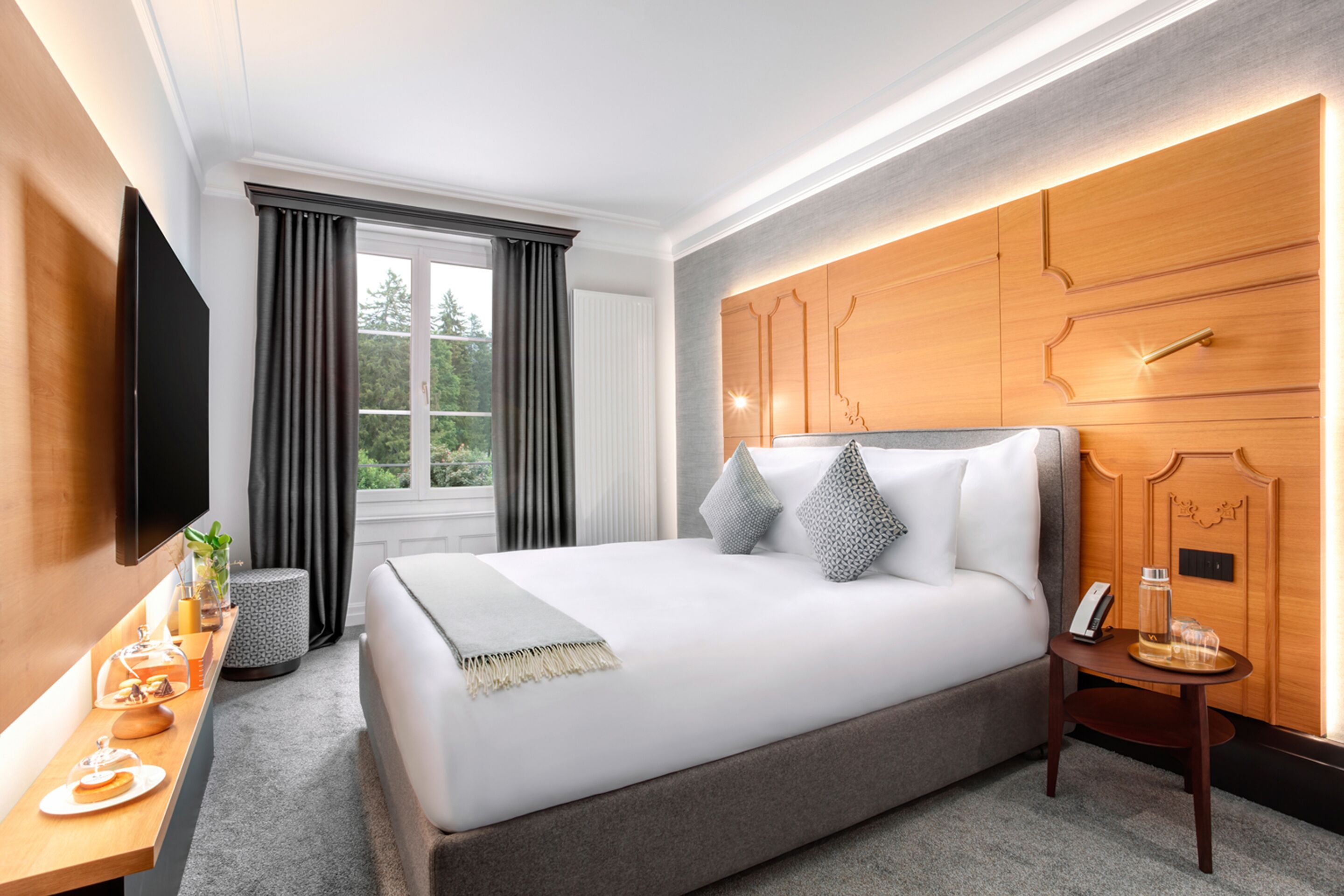 A modern hotel room with a large bed, a flat-screen TV, a wooden headboard, and a small table with a glass of water and a phone.